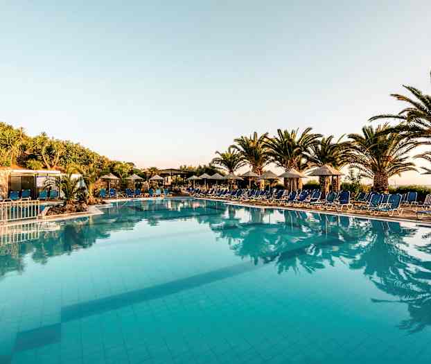 Et utendørs svømmebasseng omgitt av solsenger og parasoller, med palmer og vegetasjon i bakgrunnen. (AI-generert tekst)