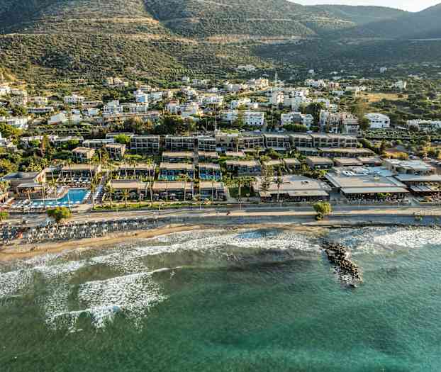 Utsikt over en strand med solsenger ved vannkanten og hotellbygninger i bakgrunnen, med grønne åser lenger bak. (AI-generert tekst)