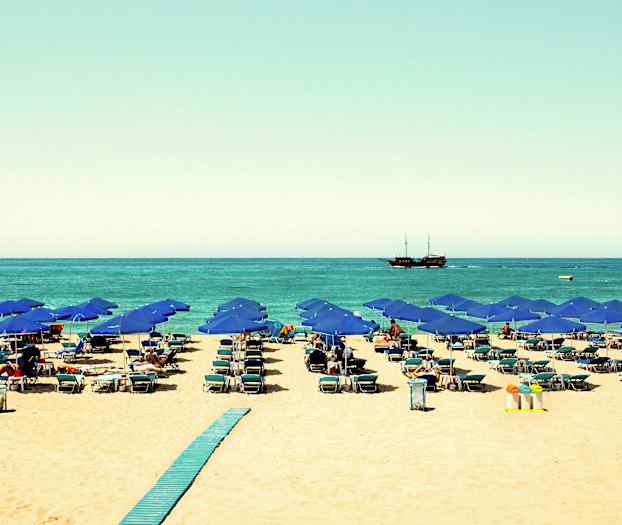 Strand med blå parasoller, solsenger og et skip på havet. (AI-generert tekst)