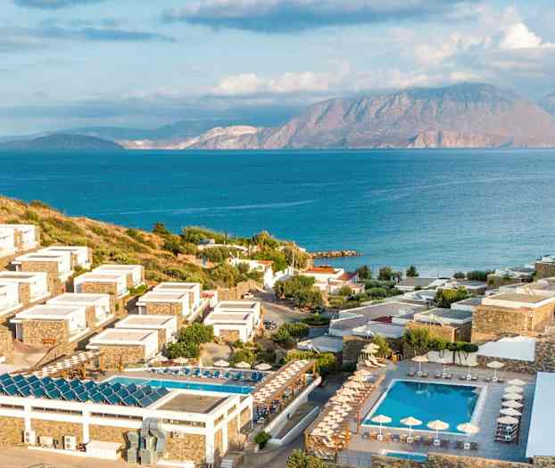 Bilde av et kystnært feriested med hotellbygninger på skrånende terreng, omgitt av grønne områder og bassenger, med hav og fjell i bakgrunnen. (AI-generert tekst)