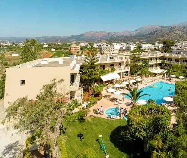 Luftbilde av hotell med basseng og hage, omgitt av grøntområder og bygninger med fjell i bakgrunnen. (AI-generert tekst)