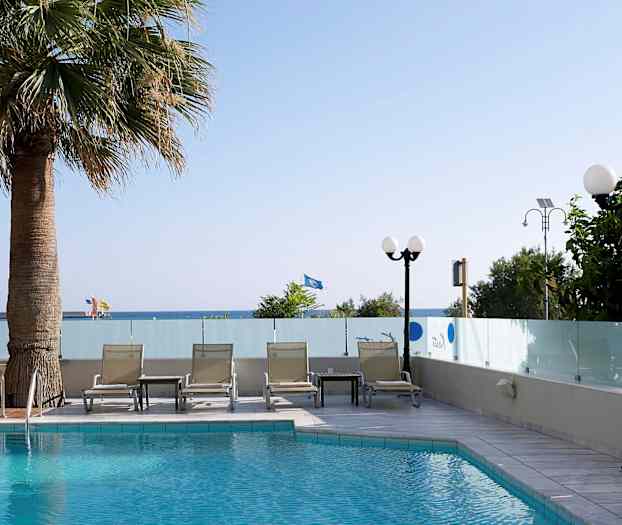 Et basseng med solsenger og et bord ved siden av, omgitt av glassvegger. Trær og en utsikt mot havet i bakgrunnen. (AI-generert tekst)