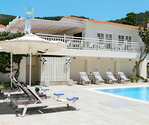 Hvit bygning med røde tak og bassengområde med solsenger og parasoller. (AI-generert tekst)