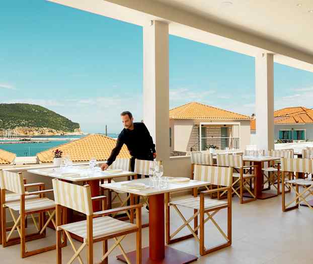 Restaurant med bordoppsett, utsikt over havet og terrasse, med bakgrunn av terrakottatak og en frodig åsside. (AI-generert tekst)