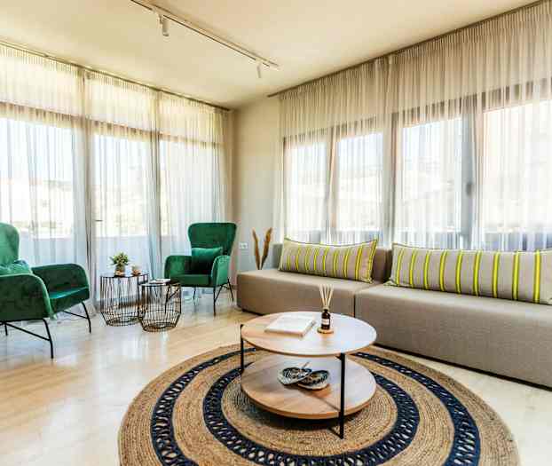 Et lyst oppholdsrom med store vinduer, to grønne lenestoler, en beige sofa med stripete puter, og et rundt salongbord med dekorasjoner. (AI-generert tekst)