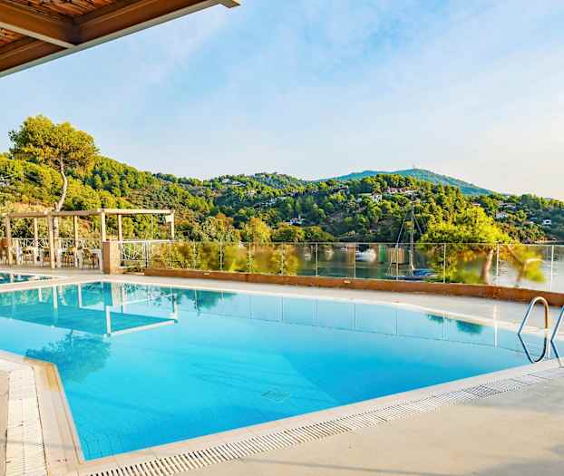 Et utendørs bassengområde med utsikt over grønne åser og en rolig innsjø. (AI-generert tekst)