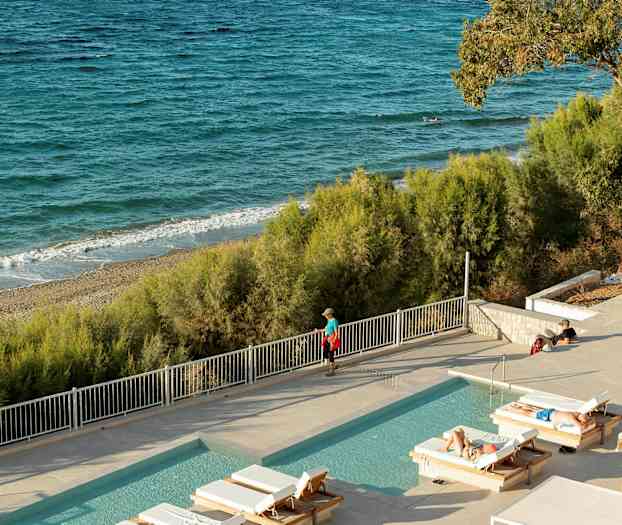 Et utendørs basseng med solsenger foran en sandstrand og havet. (AI-generert tekst)