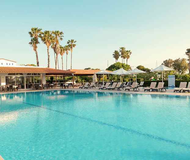 Utendørsbasseng med solsenger og parasoller ved siden av. Bak sees en bygning med skiltet 'SNACK BAR' og palmer i bakgrunnen. (AI-generert tekst)