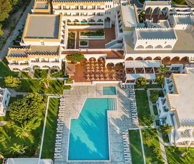 Flyfoto av et feriehotell med bassengområde og omkringliggende bygninger. (AI-generert tekst)