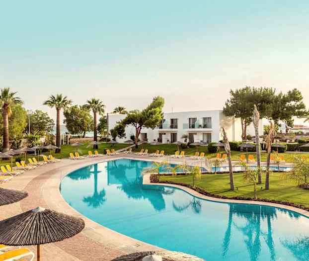 Stor basseng omgitt av solsenger og parasoller, med grønne plener og palmer i bakgrunnen samt hvite bygninger. (AI-generert tekst)