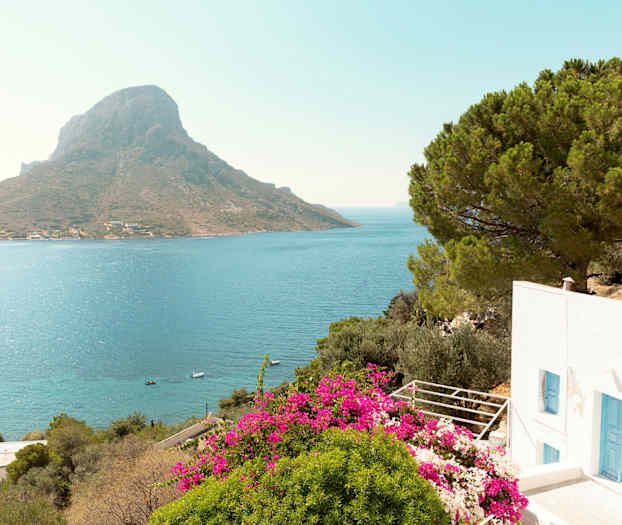 En hvit bygning med blå dører, omgitt av grønt og rosa blomster, med utsikt over havet og et fjell i bakgrunnen. (AI-generert tekst)