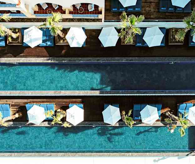 Et fugleperspektivbilde av to parallelle svømmebassenger omgitt av palmer, solsenger og parasoller. (AI-generert tekst)