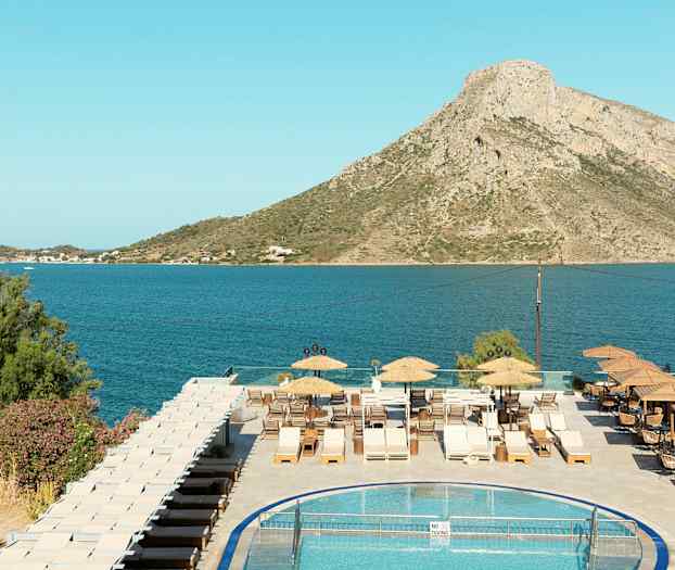 Utsikt over et basseng, en terrasse med solsenger, og en sjøutsikt med fjell i bakgrunnen. (AI-generert tekst)