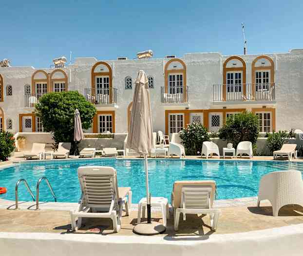 Et hotellbasseng med solsenger og parasoller foran en bygning med hvite fasader og oransje detaljer. (AI-generert tekst)