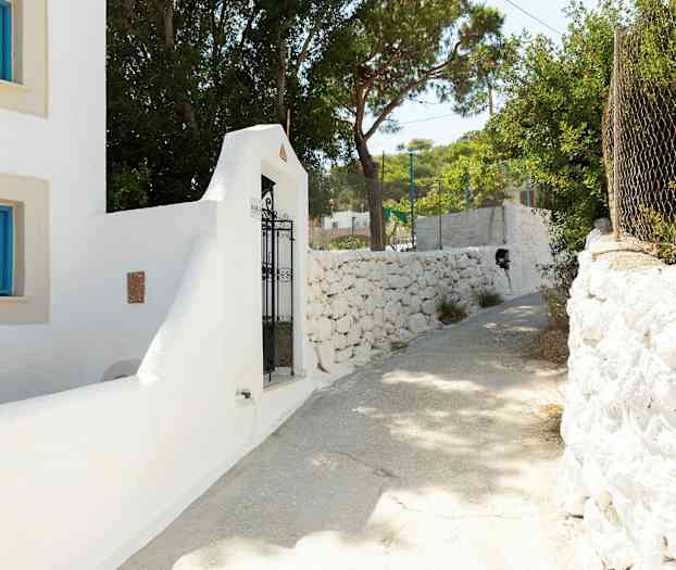 Hvit bygning med blå vinduskarmer og en smijernsport ved en steinmur. Solrik gangvei omgitt av trær. (AI-generert tekst)