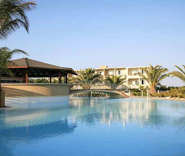 Utendørs svømmebasseng med bro, omgitt av palmer og en bygning i bakgrunnen. (AI-generert tekst)