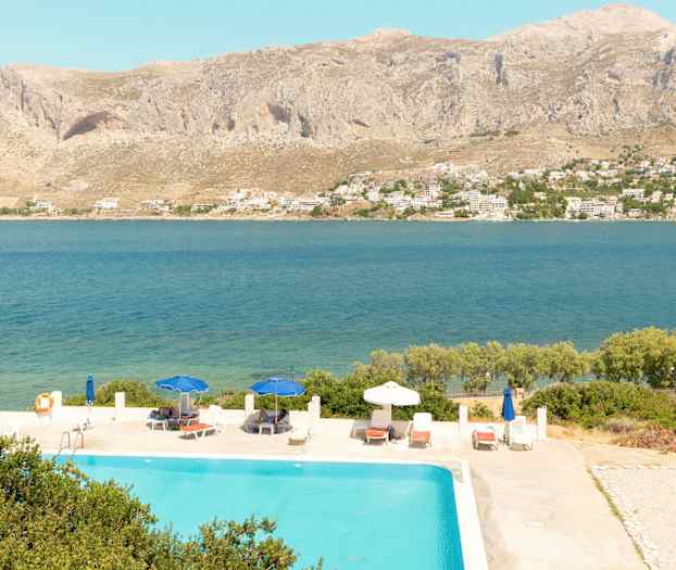 Utsikt over et basseng med solsenger og parasoller, med havet og fjell i bakgrunnen. (AI-generert tekst)