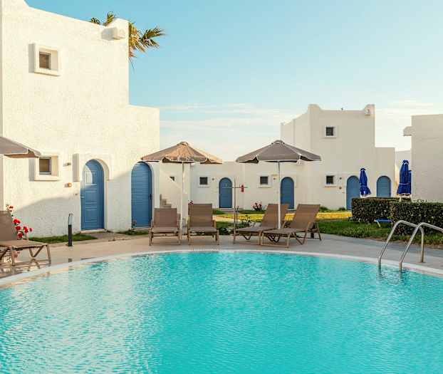 Et basseng omgitt av solsenger og parasoller, med hvite bygninger med blå dører i bakgrunnen. (AI-generert tekst)