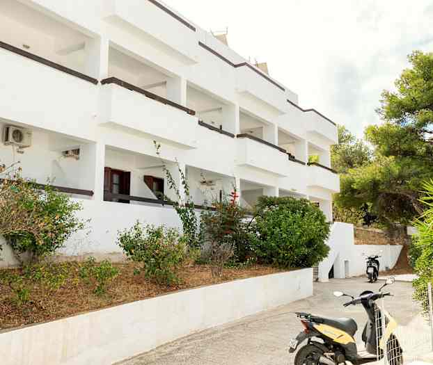 Hvit bygning med balkonger omgitt av busker og en parkert motorsykkel. (AI-generert tekst)