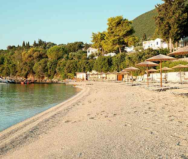 En strand med solstoler og parasoller, vegetasjon og bygninger i bakgrunnen. (AI-generert tekst)