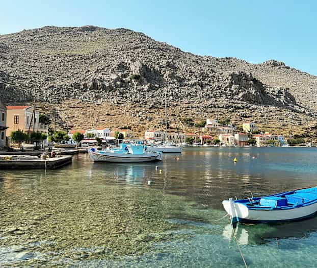 En havn med klart vann, små båter og fargerike hus ved foten av steinete fjell. (AI-generert tekst)