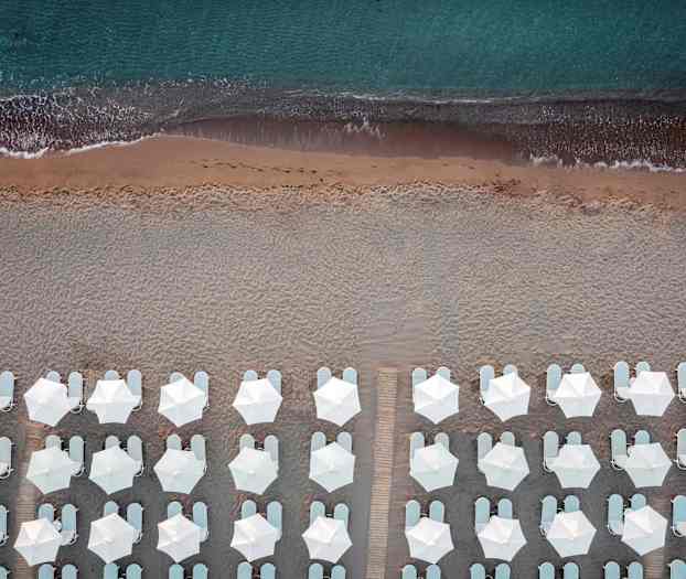 Fugleperspektiv av strand med hvite parasoller og solsenger ved kanten av havet. (AI-generert tekst)