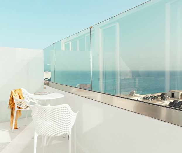 En hvit balkong med to stoler og et bord, med utsikt over havet og bygninger. (AI-generert tekst)