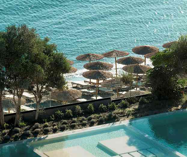 Utsikt over basseng, trær og stranden med parasoller og solsenger mot sjøen. (AI-generert tekst)