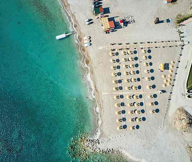 Luftfoto av en strand med parasoller og solsenger ved siden av klart blått vann. (AI-generert tekst)