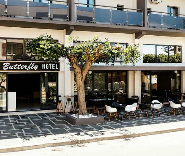 Forsiden av et hotell med skiltet "Butterfly Hotel", en uteplass med bord og stoler samt et tre foran bygningen. (AI-generert tekst)