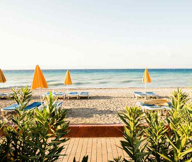 En strand med solsenger og gule parasoller ved et blått hav, omgitt av planter. (AI-generert tekst)