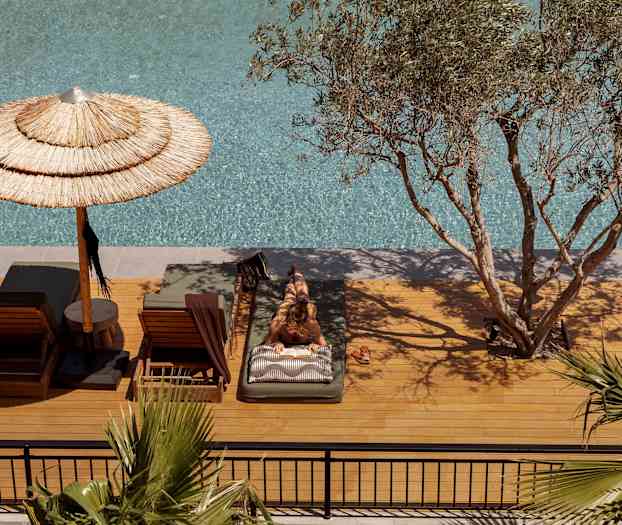 Person ligger på en solseng ved et basseng, omgitt av trær og parasoller. (AI-generert tekst)