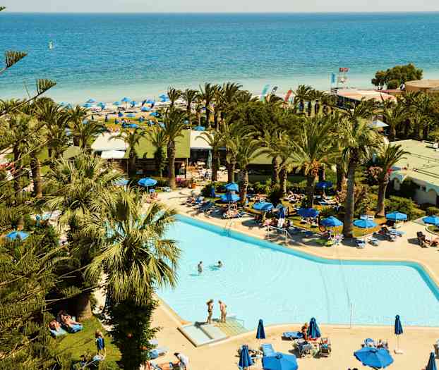 Et svømmebasseng omgitt av solsenger og palmer, med en sandstrand og havet i bakgrunnen. (AI-generert tekst)