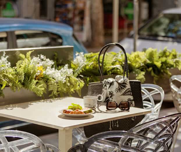 Bord med croissant, kopp og solbriller på uteservering med blomsterdekorasjoner. (AI-generert tekst)