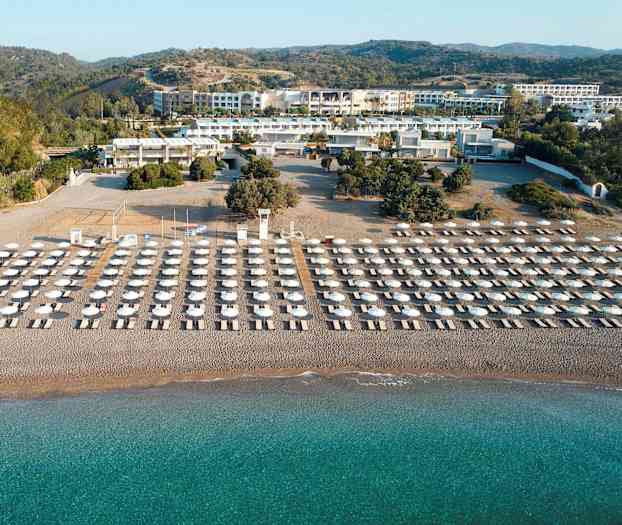 Solstoler og parasoller i flere rader på en sandstrand med turkis vann, omgitt av grøntområder og bygninger i bakgrunnen. (AI-generert tekst)