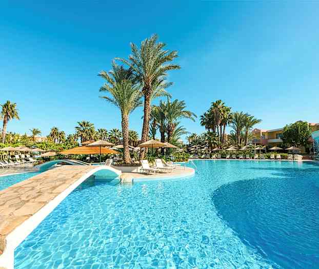 Et stort utendørs basseng med en liten bro over, omgitt av solsenger og palmer. (AI-generert tekst)