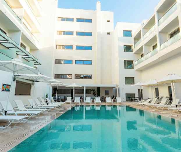 Bilde av et hotellområde med et svømmebasseng omgitt av solsenger og parasoller ovenfor. Bakgrunnen viser hvite bygninger med balkonger. (AI-generert tekst)