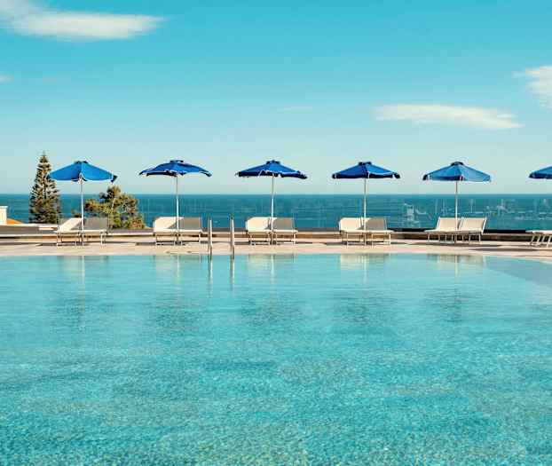 Utendørsbasseng med blå parasoller og utsikt over havet. (AI-generert tekst)