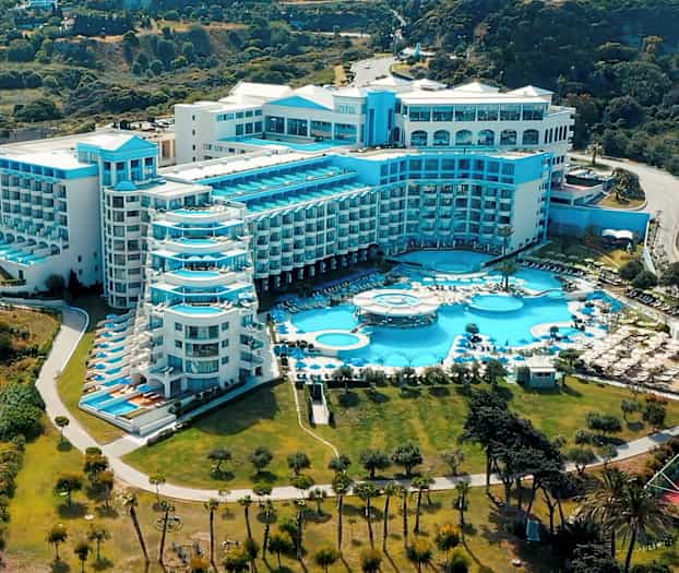 Stort hotellbygg med flere blå balkonger og bassenger, omgitt av grøntområder. (AI-generert tekst)