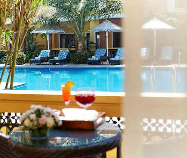 Bildet viser et basseng med solsenger og parasoller samt et nærbilde av et bord med drikke og blomster i forgrunnen. (AI-generert tekst)