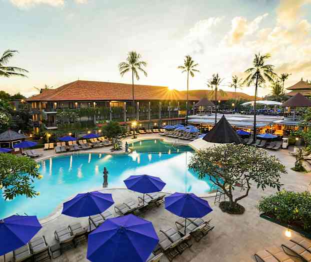 Bilde av en resort med basseng, omgitt av solstoler og parasoller samt grønne palmer i bakgrunnen. (AI-generert tekst)