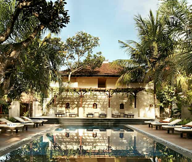 Et utendørs basseng omgitt av solsenger og trær med en bygning i bakgrunnen. (AI-generert tekst)