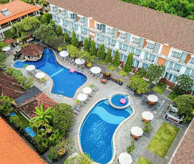 Et fugleperspektiv av et hotellområde med to bassenger, solsenger og parasoller, omgitt av vegetasjon og bygninger. (AI-generert tekst)