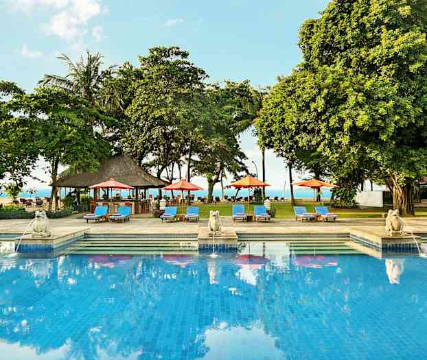 Et utendørs svømmebasseng omgitt av solsenger, parasoller og trær med havet i bakgrunnen. (AI-generert tekst)