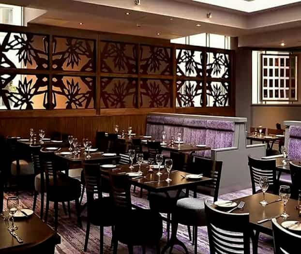 En stilfull restaurant med bord og stoler oppstilt, en dekorativ trevegg med blomstermønster og lilla detaljer på sitteplasser. (AI-generert tekst)
