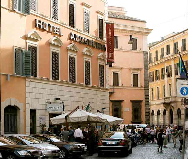 En gatescene med en hotellbygning med beige fasade og mørke vinduslemmer. Foran bygningen står flere parkede biler og en kafé med parasoller. (AI-generert tekst)