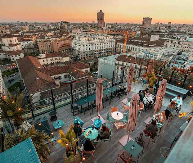 En takterrasse med bord og sitteplasser, omgitt av panoramautsikt over en by ved solnedgang. (AI-generert tekst)