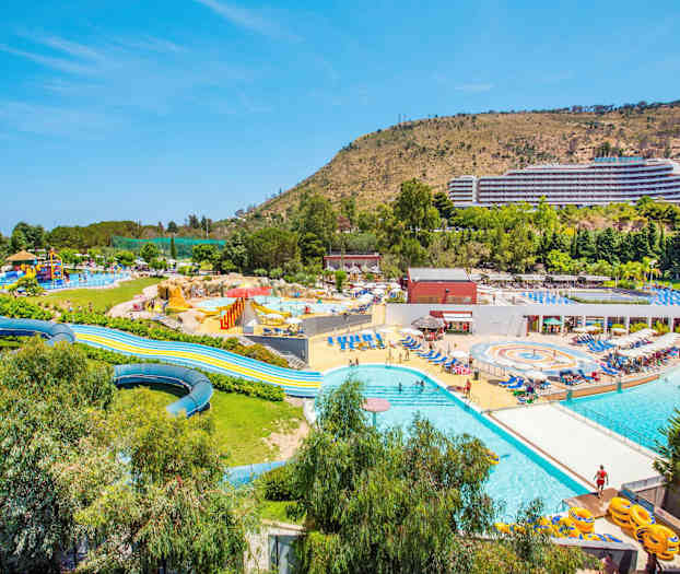 Bildet viser et stort utendørs badeland med bassenger, vannsklier og omkringliggende solsenger. En stor bygning og skrånende terreng er synlig i bakgrunnen. (AI-generert tekst)