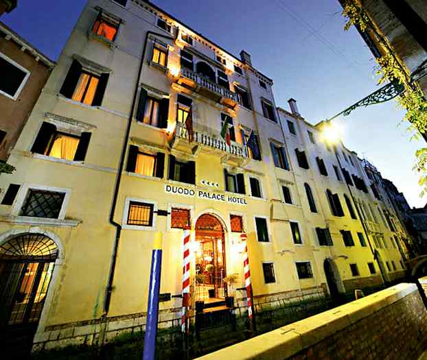 Leilighetsbygning i tre etasjer med skilt Duodo Palace Hotel, omgitt av en kanal og en gangvei. (AI-generert tekst)