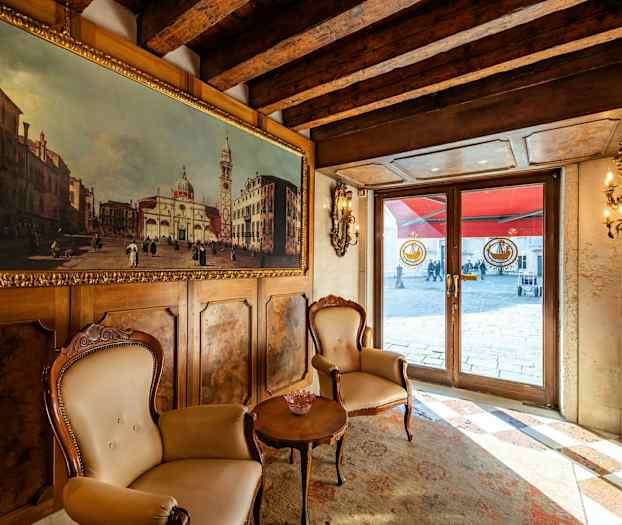 Innendørs med to stoler og et bord foran en vegg med et maleri, med utsikt mot en utvendig gjennom glassdører. (AI-generert tekst)